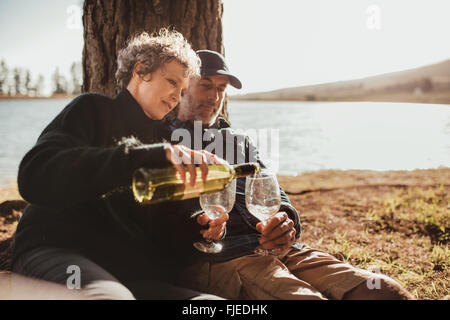 Senior man and woman enjoying outdoors camping on picnic area near Lake. Woman pouring wine in glasses, both sitting under a tre Stock Photo