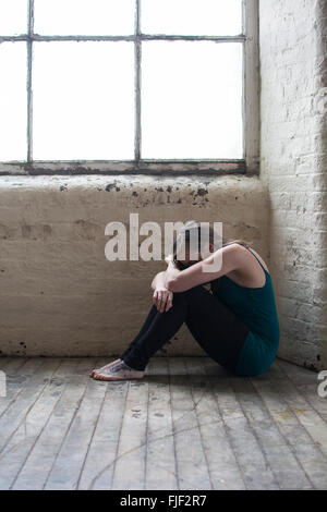 Sad young woman sat on the wooden floor by the window Stock Photo