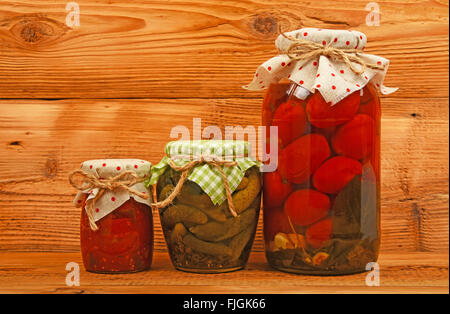 Three glass jars of homemade conserves, eggplant pepper salad, pickled tomatoes and cucumbers with textile decoration over brown Stock Photo