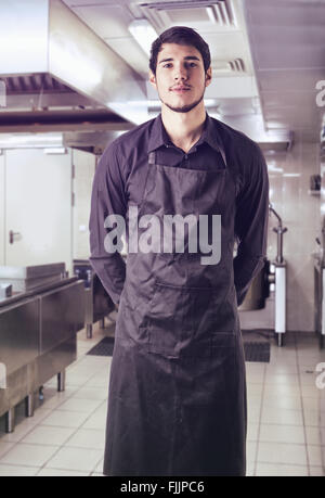Young chef or waiter posing, wearing black apron and shirt standing in restaurant kitchen Stock Photo