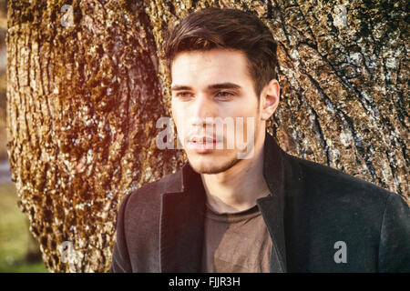 Handsome young man leaning against tree, looking away to a side, in a sunny day Stock Photo