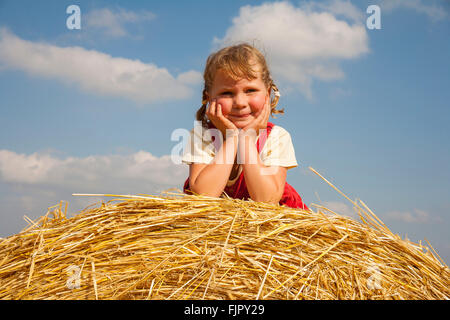 Little girl lying on a straw bale under a cloudy blue sky, Germany Stock Photo