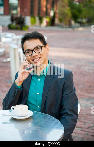 Successful young asian man in business casual attire sitting and smiling in relaxing outdoor cafe with cup of coffee talking on Stock Photo