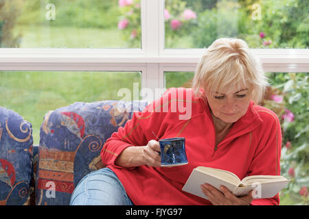Blonde middle-aged woman is sitting  on the sofa and and reading a book. Woman is holding a blue cup. Stock Photo