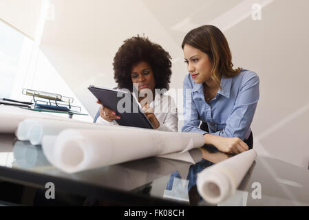 Two architects in modern office building, sitting on desk with blueprints and housing projects. The women hold a tablet and surf Stock Photo