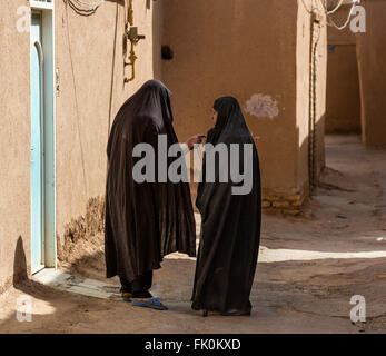 Two chador-clad women engaging in animated conversation in an alley lined with mud brick houses in Yazd, Iran Stock Photo