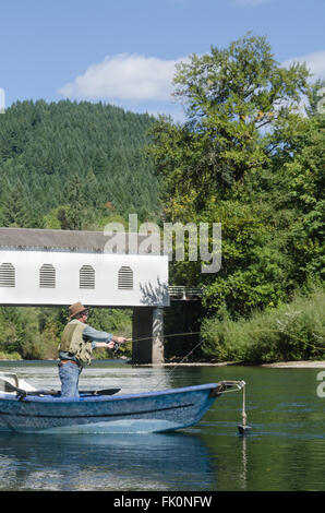 A fisherman in a drift boat casts his line just below the Goodpasture covered bridge on the Mckenzie River near Eugene Oregon Stock Photo