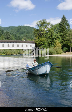 A fisherman in a drift boat rows just below the Goodpasture covered bridge on the Mckenzie River near Eugene Oregon Stock Photo