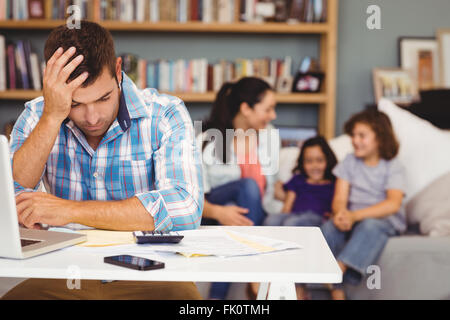 Tensed man by laptop while family sitting in background Stock Photo