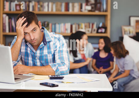 Tensed man by laptop table while family sitting in background Stock Photo