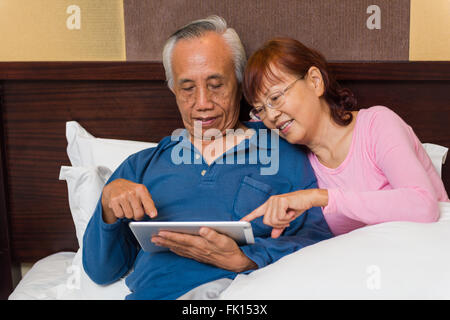 Asian senior couple relaxing and learning new technology on mobile device in bed. Stock Photo