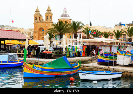 Traditional Maltese luzzu fishing boats in Marsaxlokk harbour with the church of Our Lady of Pompeii behind, Marsaxlokk, Malta. Stock Photo