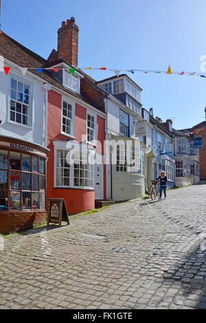 Lymington Hampshire colorful rendered walls and bay windows on shop fronts along hilly cobbled Quay Hill street woman walking with bike England UK Stock Photo