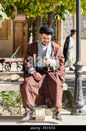 Mullah checks his mobile phone while sitting on a bench at the Khan Madrassa, Shiraz, Iran Stock Photo