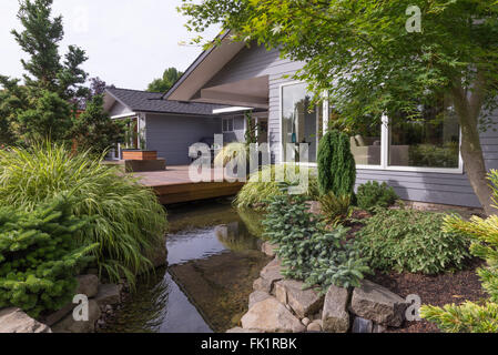 A water feature emulating a creek flows under a deck and between landscaped rocks with a contemporary home in the background. Stock Photo