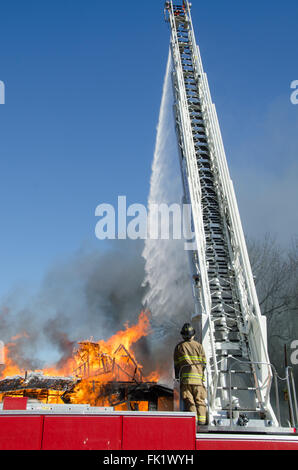 A fireman standing at the controls of an105 foot reach aerial ladder/truck directs a sream of water on a blazing house. Stock Photo