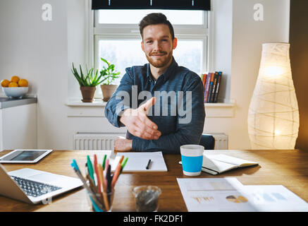 Smiling businessman leaning across his desk offering his hand in greeting, to close a deal, in partnership or congratulations Stock Photo
