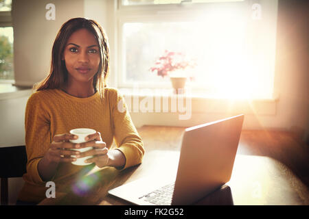 Serious young single female sitting at table holding coffee cup next to open laptop with bright sun coming through window Stock Photo