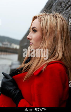 Profile of a young woman wearing a red coat leaning against an old stone wall Stock Photo