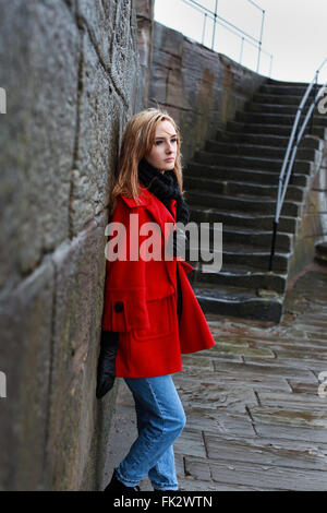 Woman in a red coat leaning on an old stone wall in the sleet and rain Stock Photo