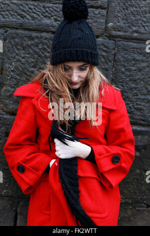 Woman wearing a red jacket and pom pom hat leaning on an old stone wall in the sleet and rain Stock Photo