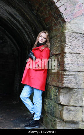 Woman wearing a red coat leaning against an old wall of a tunnel Stock Photo