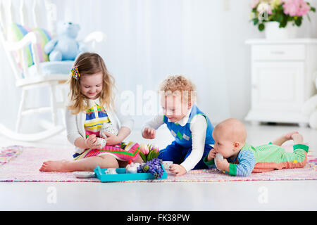 Kids play at home. Children playing in white sunny nursery. Little girl having fun at doll tea party with her brothers. Siblings Stock Photo