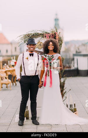 Cheerful african newlyweds holding hands and smiling at the wedding ceremony on the rooftop Stock Photo