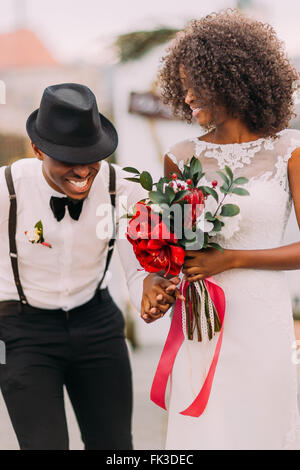Stylish black groom in hat and his charming bride happily laughing on their wedding ceremony Stock Photo