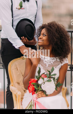 Happy african bride and groom with bouquet of red flowers softly holding hands Stock Photo