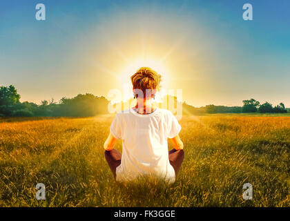 Back view of young adult blonde woman sitting in the field meditating in front of the sun. Stock Photo