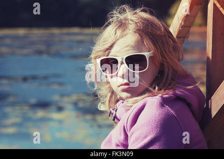 Blond teenage girl in sunglasses sitting on a river coast, outdoor closeup portrait with tonal correction photo filter effect, i Stock Photo