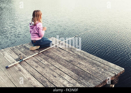 Little blond Caucasian girl sitting on a wooden pier with fishing rod, vintage stylized photo with warm tonal correction filter Stock Photo