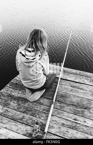 Little blond Caucasian girl sitting on a wooden pier with fishing rod, black and white photo, vertical composition Stock Photo