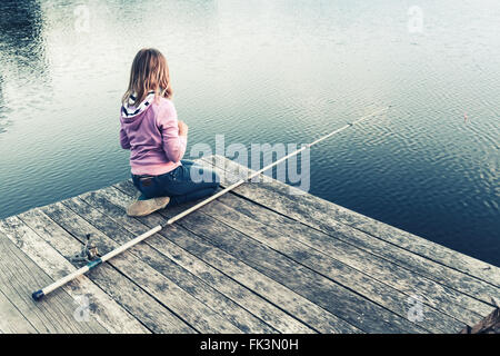 Little blond Caucasian girl sitting on a wooden pier with fishing rod, vintage stylized photo with tonal correction filter effec Stock Photo