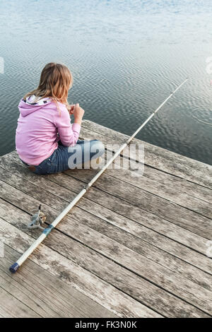 Little blond Caucasian girl sitting on a wooden pier with fishing rod Stock Photo