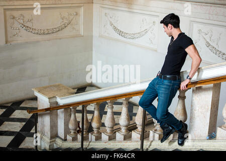 Side view of young dark haired man going down on railing in museum. White walls decorated with ornament. Stock Photo