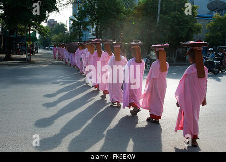 Long line of Buddhist nuns dressed in pink robes, heads covered, alms bowls on their heads, crossing main road in Mandalay Stock Photo