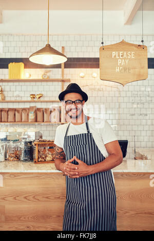 Coffee shop worker smiling to camera, standing at the counter. Happy young man in apron and hat leaning to cafe counter. Stock Photo