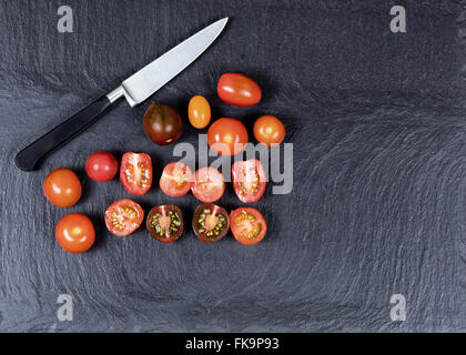 Overhead view of colorful mixed tomatoes, whole and sliced, with knife on natural black slate stone. Stock Photo