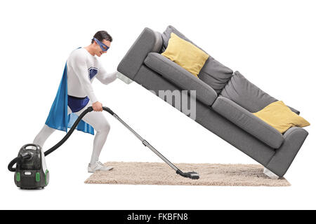 Profile shot of a strong man in superhero costume vacuuming under a sofa isolated on white background Stock Photo