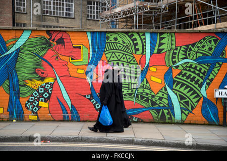 Hanbury Street, off Brick Lane. Tower Hamlets. Two women wearing black coats and veils walk past a colorful mural. Stock Photo