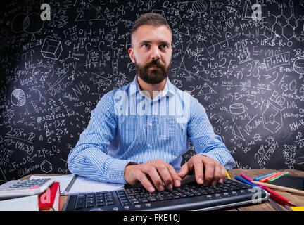 Happy teacher sitting at desk, school supplies, big blackboard Stock Photo