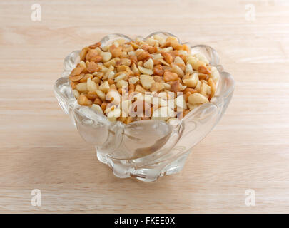 Side view of roasted and salted macadamia nut pieces in a glass bowl on a wood table top illuminated with natural light. Stock Photo