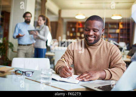 Young businessman writing plans in his notebook with business people behind him Stock Photo