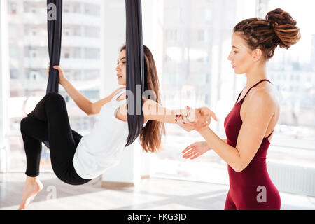 Serious young woman trainer teaching beautiful girl doing aerial yoga in studio Stock Photo