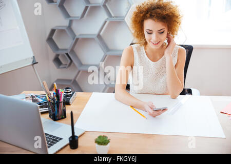 Happy curly pretty young woman photographer sittting and using smartphone in the office Stock Photo