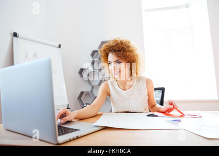 Cheerful curly redhead young woman cutting color paper and using laptop in office Stock Photo