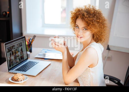 Beautiful smiling redhead young woman drinking coffee at the table and using laptop in the office Stock Photo