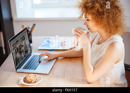 Profile of attractive charming curly young woman photographer drinking coffee and working with laptop on workplace Stock Photo